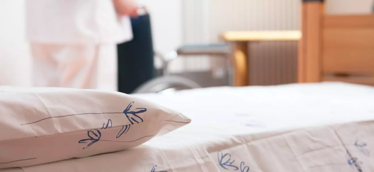 Bed at nursing home with nurse and wheelchair on background.