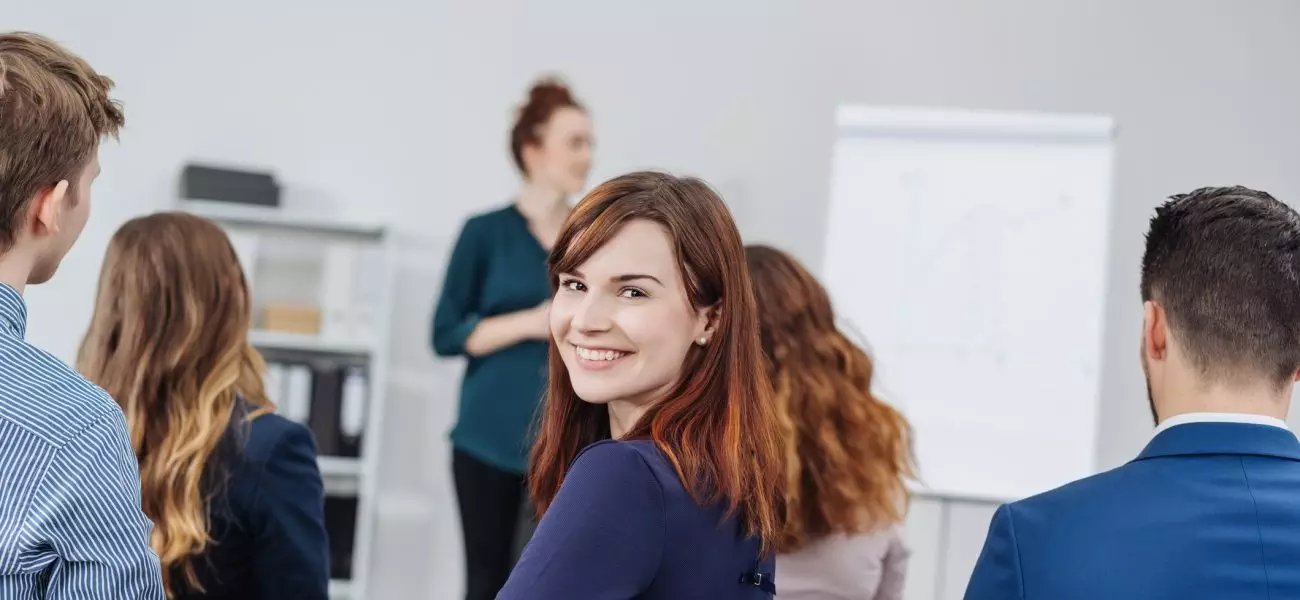 A smiling, happy woman turns in her chair to look at the camera whilst surrounded by other students during a lecture in a classroom.